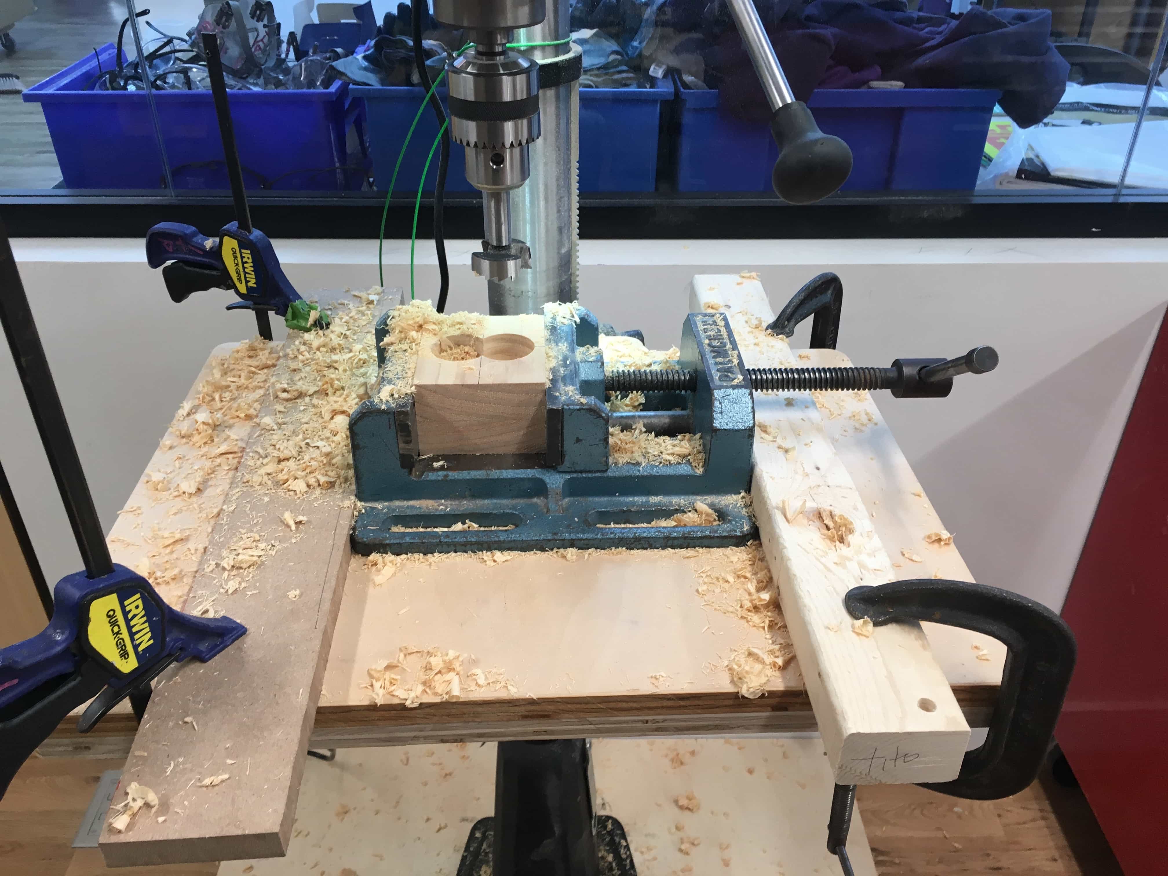 Block of wood in clamp, secured to drill press. Two holes have been drilled.